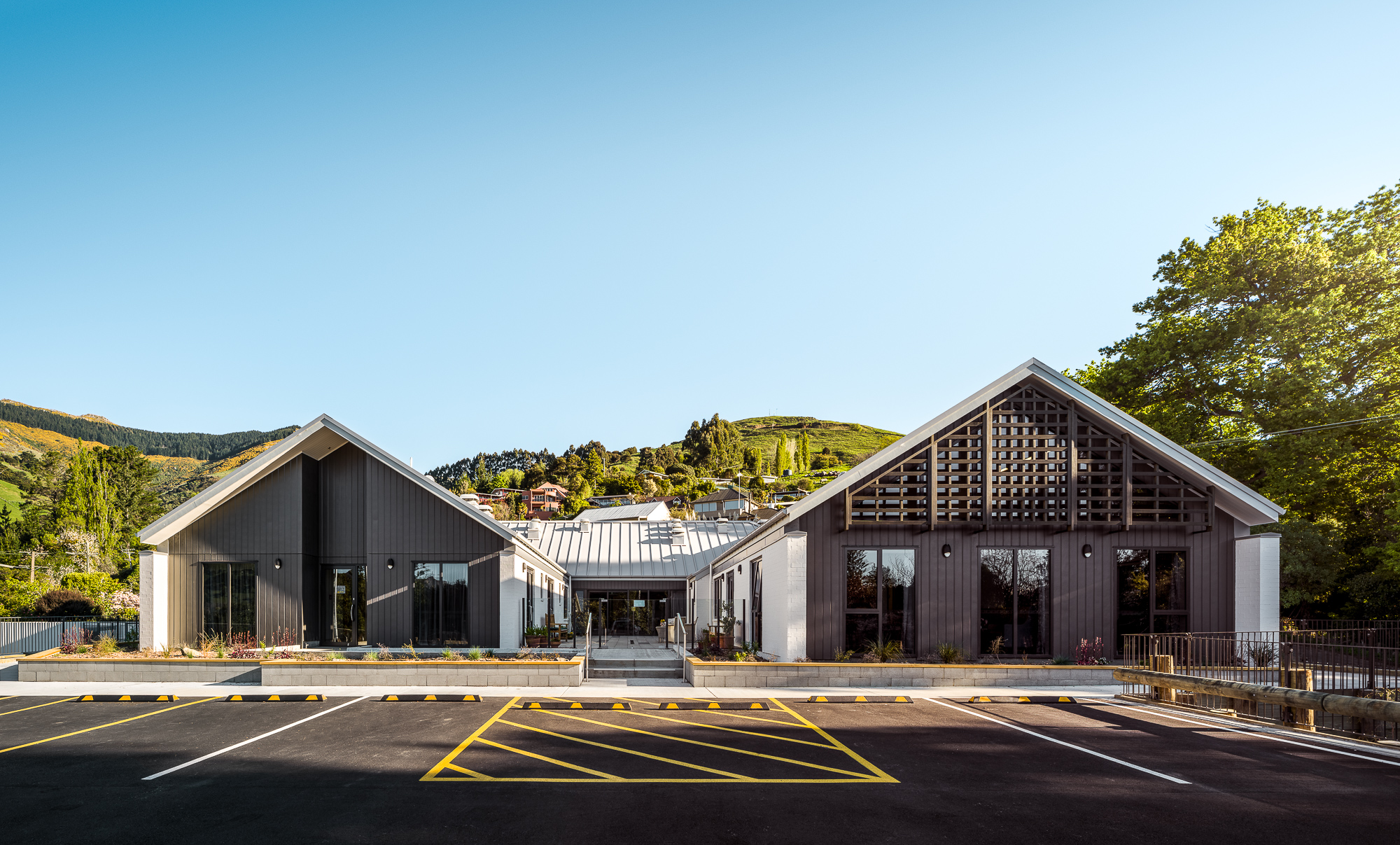Front elevation of the Akaroa Health Hub showing twin gable forms, large glazing, and accessible pathways, with green hills in the background.