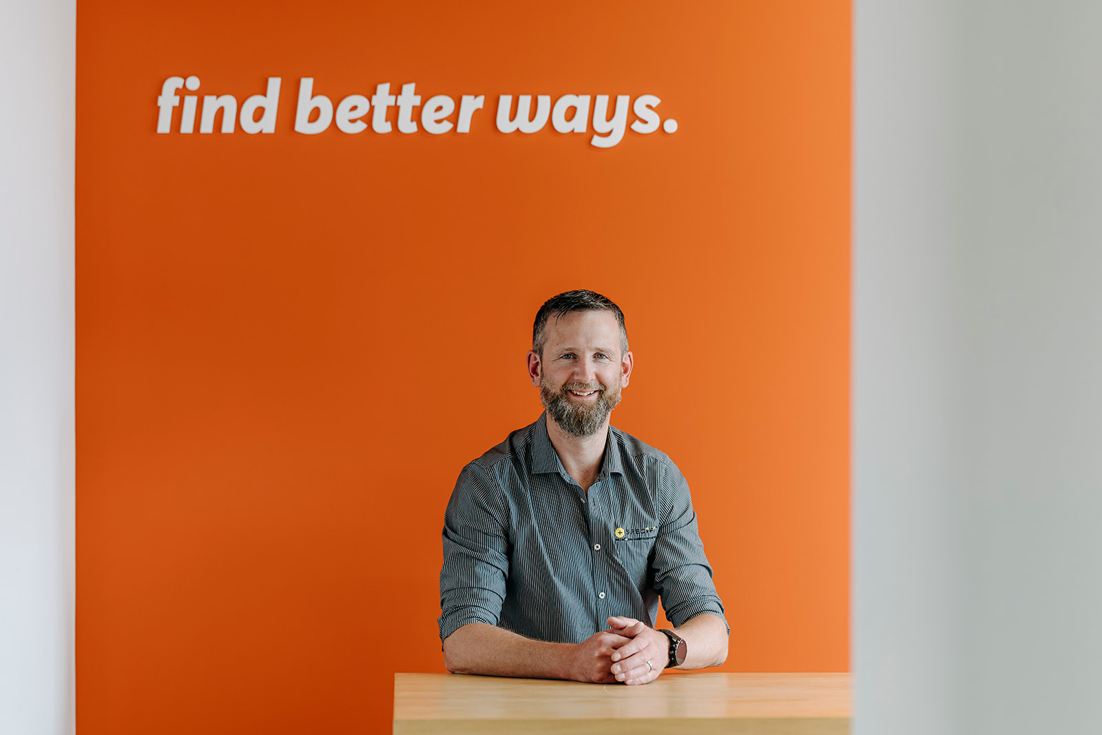Aidan Hynes, Specifi Manager at Kirk Roberts, seated in front of an orange wall featuring the company tagline “Find better ways.”